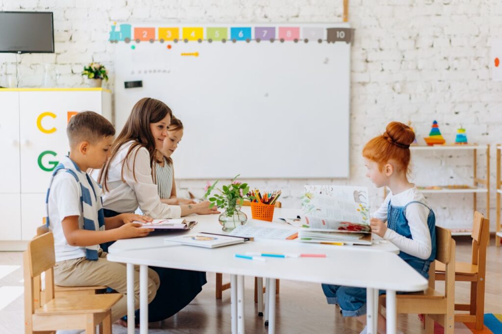 SEND_Education_Help_Images - Northants Send Family Guidance Adult teacher and three children sit at a white table, looking at books and worksheets in a bright classroom with a whiteboard and colorful decor in the background