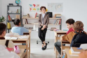 SEND_Education_Support_Images - Northants Send Family Guidance Teacher standing at the front of a classroom, addressing students seated at desks with world maps and books on shelves behind.
