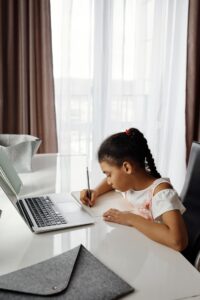 SEND_Guidance_Help - Northants Send Family Guidance Young girl with braided hair writes in a notebook at a white desk, laptop open beside her, backlit by sheer curtains.