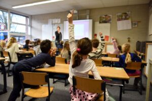 SEND_School_Help_Images - Northants Send Family Guidance A young student raises her hand in a classroom as a teacher stands at the front.