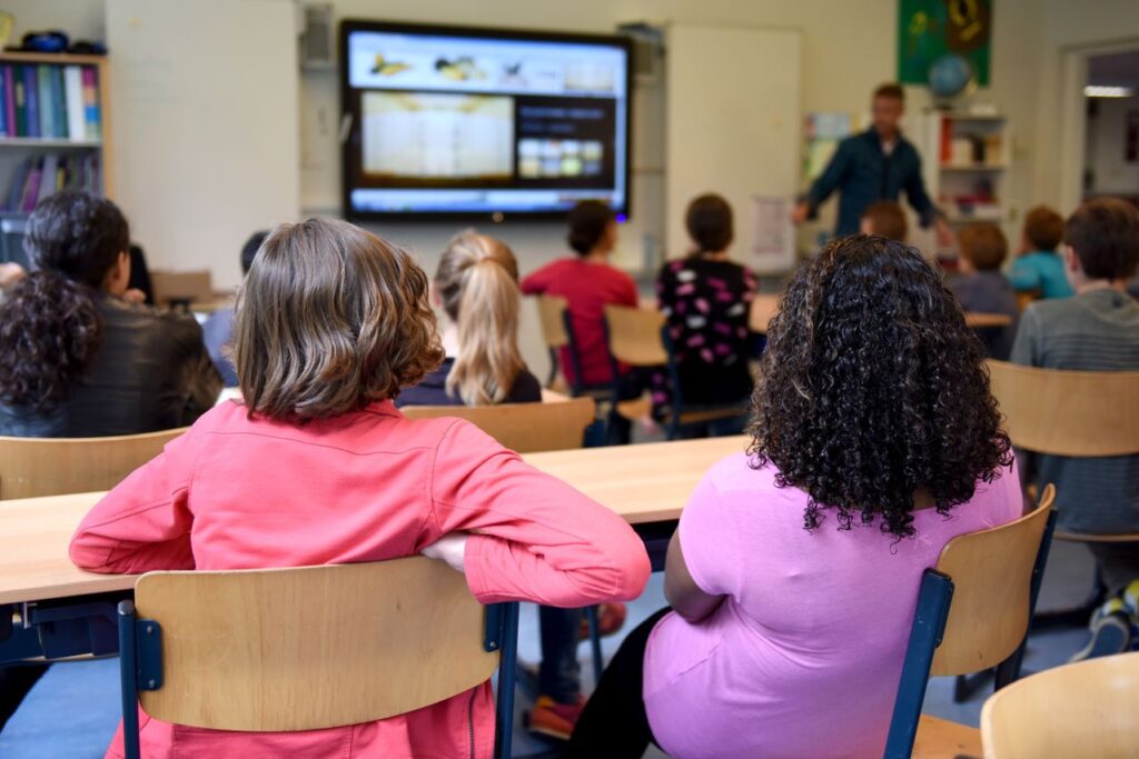 SEND_School_Images - Northants Send Family Guidance Classroom scene with students seated at desks watching a teacher present on a screen at the front of the room (two girls in pink and purple in foreground).