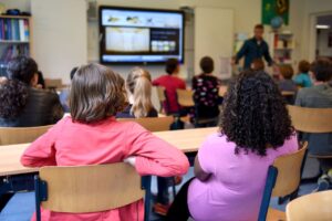 SEND_School_Images - Northants Send Family Guidance Classroom scene with students seated at desks watching a teacher present on a screen at the front of the room (two girls in pink and purple in foreground).